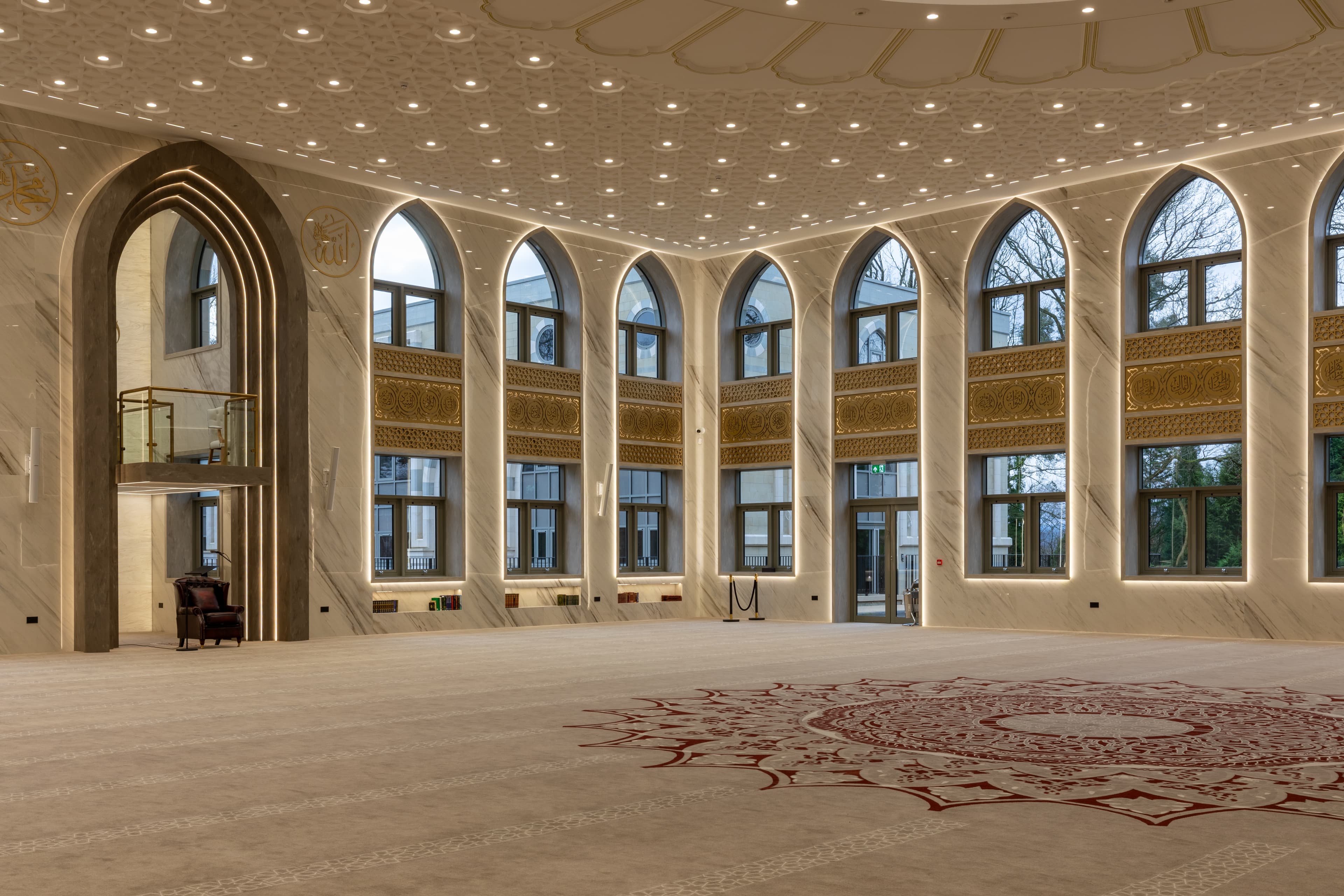 The main prayer hall in daylight, with tall arched windows curving around the carpet medallion