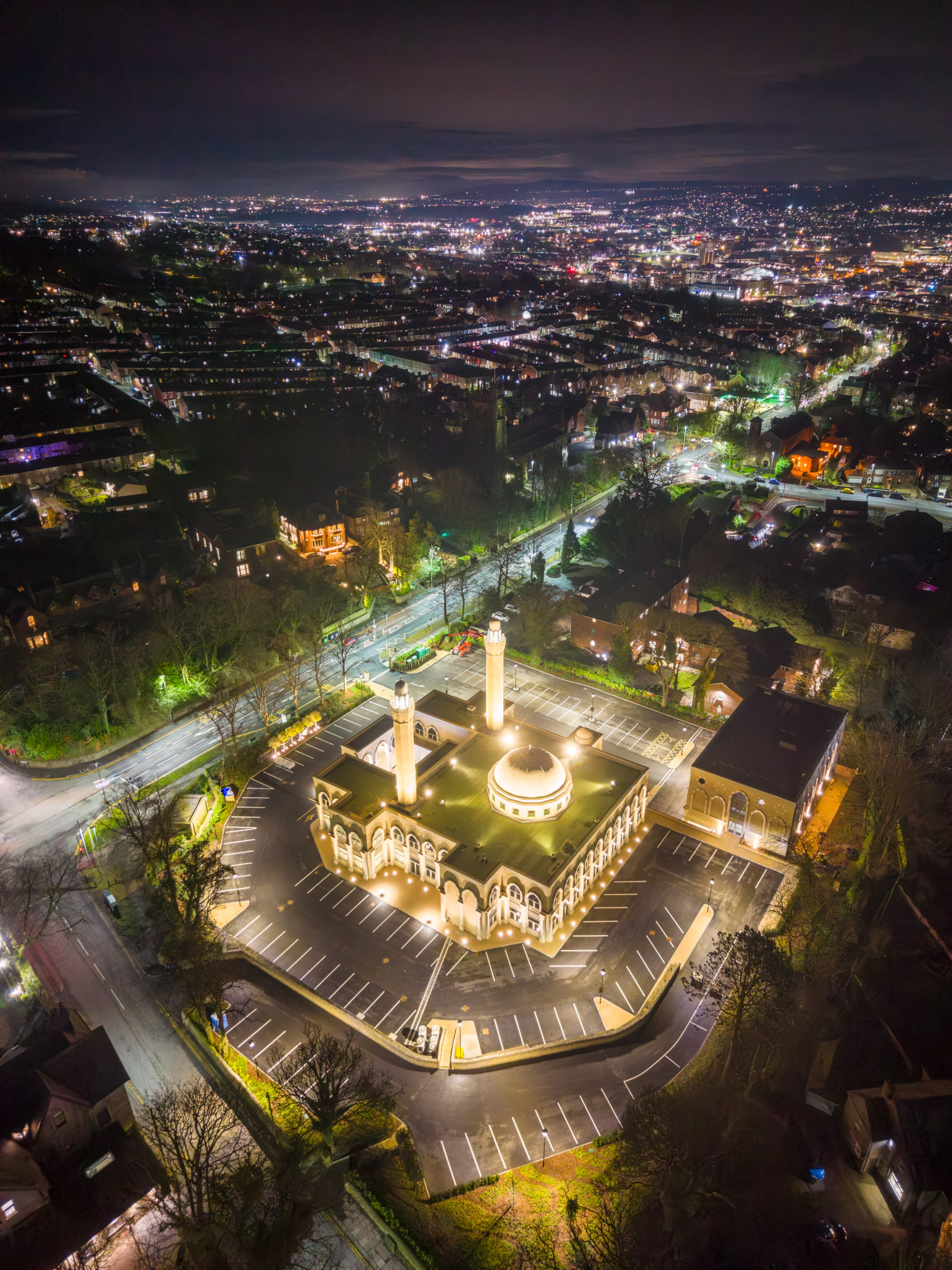 Aerial view at night of the lit mosque complex set among the dark rows of Blackburn terraced housing and street lighting.